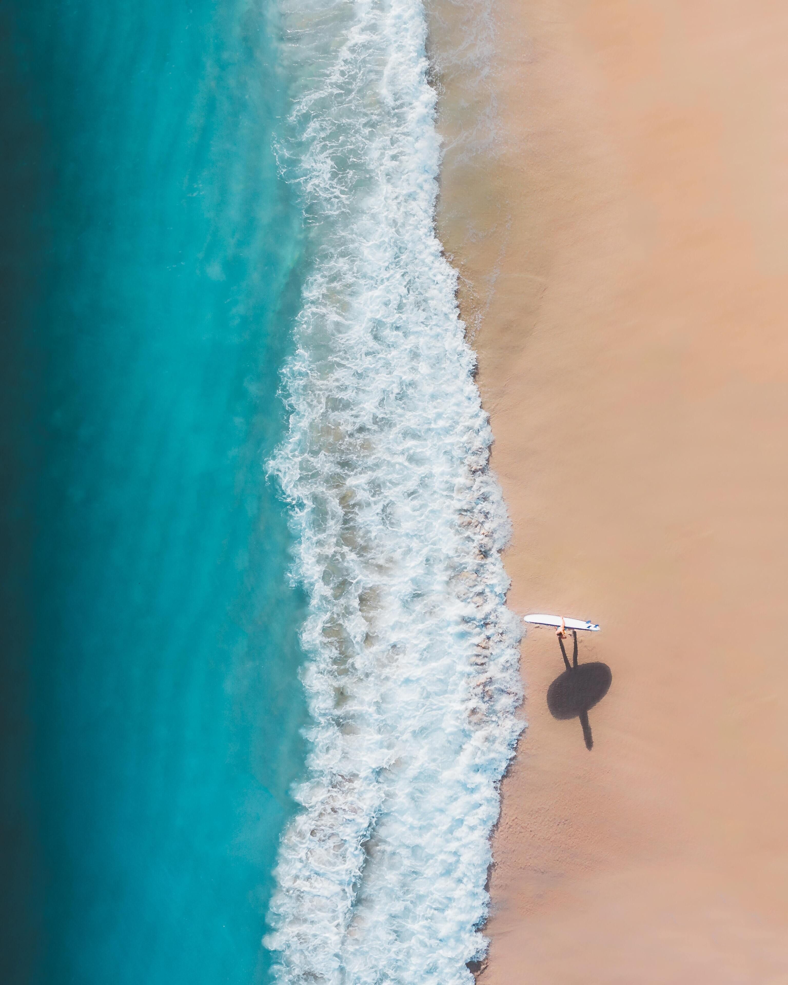 An aerial view of a beach with waves crashing onto the shore and a person holding a surfboard.