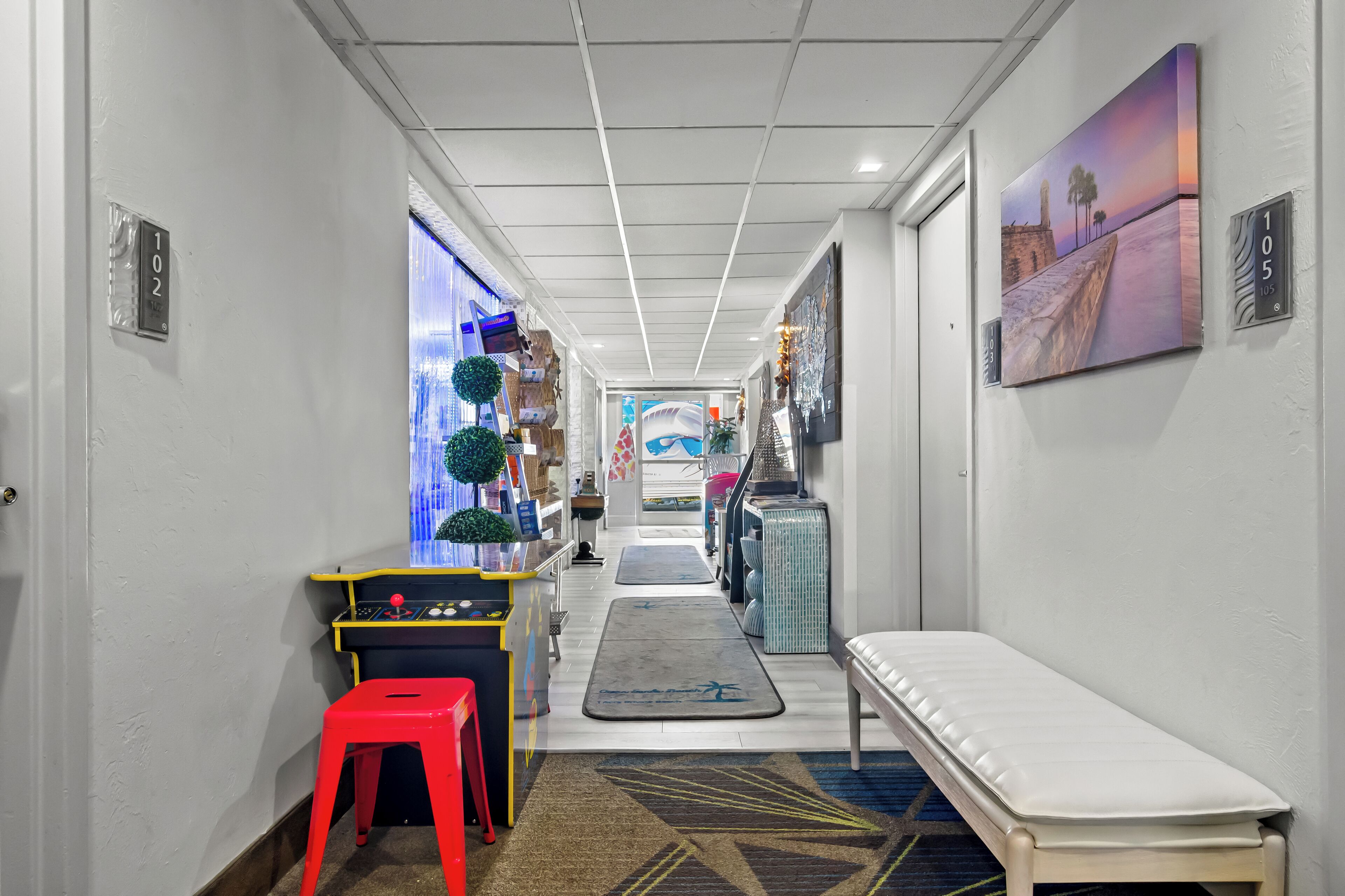 A hotel hallway with a bench, a red stool, a control panel, and a painting on the wall.