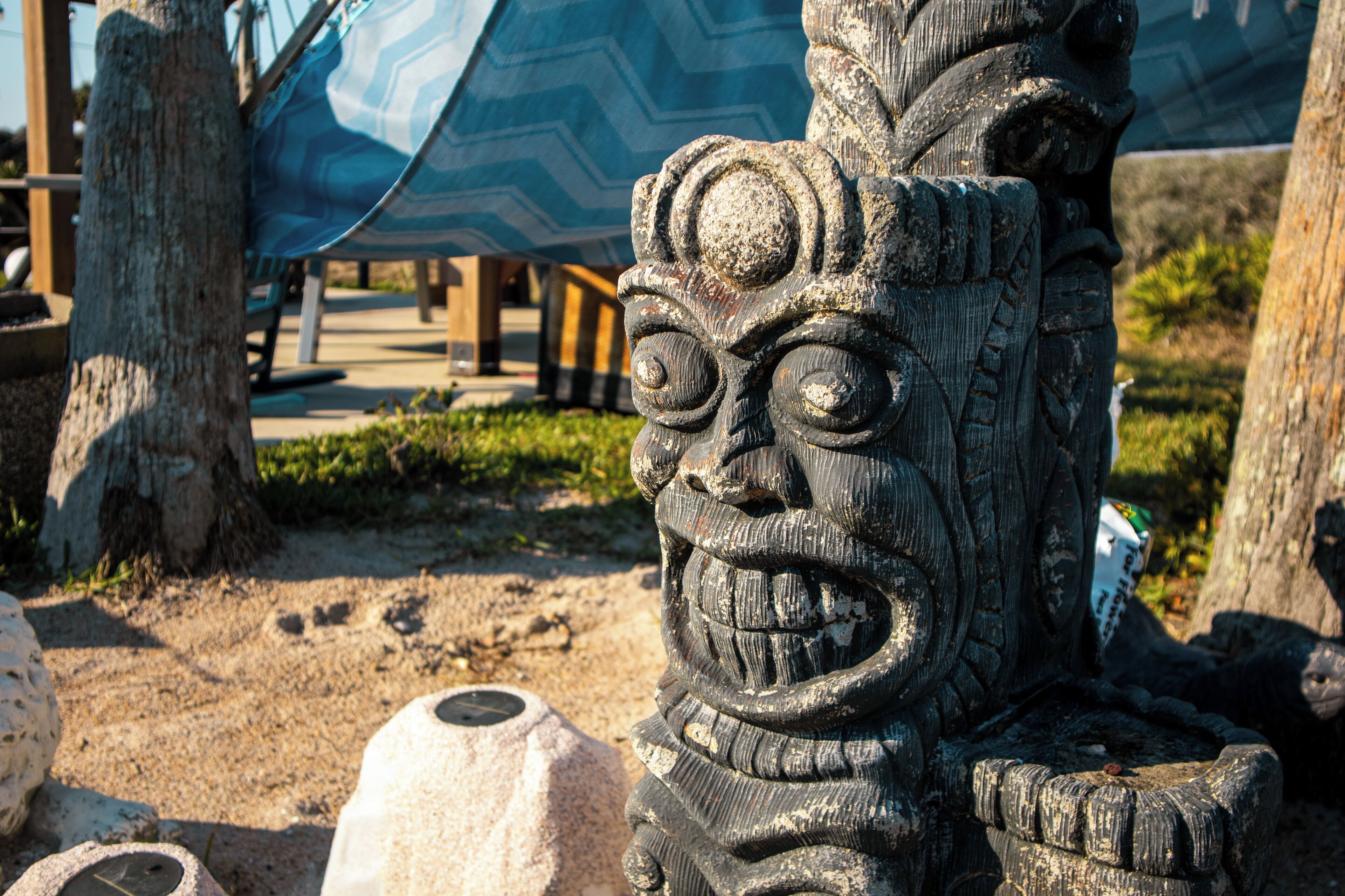 A stone statue with a carved face, surrounded by trees and a patterned fabric structure in the background.