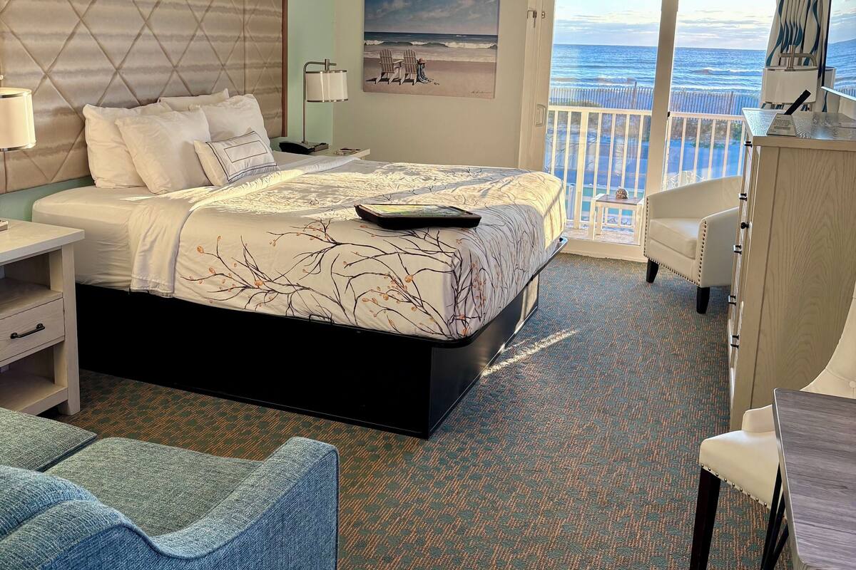 Quarto de hotel com cama, escrivaninha, cadeira e vista para o oceano.