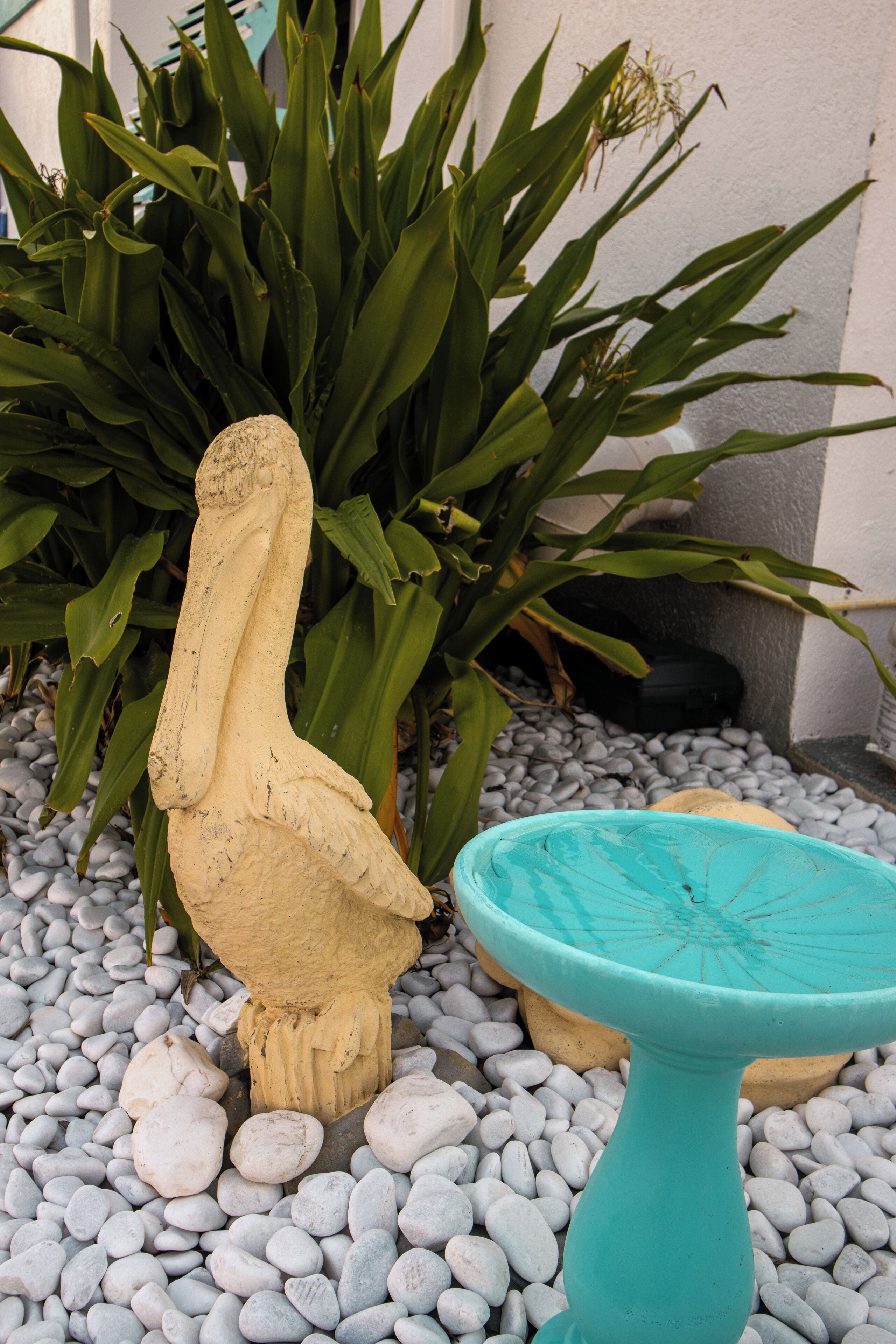 A garden with a bird sculpture and a turquoise birdbath surrounded by pebbles and green foliage.