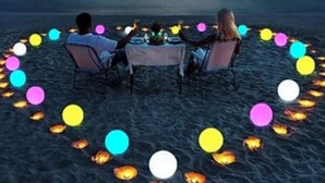 Vista al atardecer con una mesa y sillas dispuestas en la playa, rodeadas de luces flotantes.