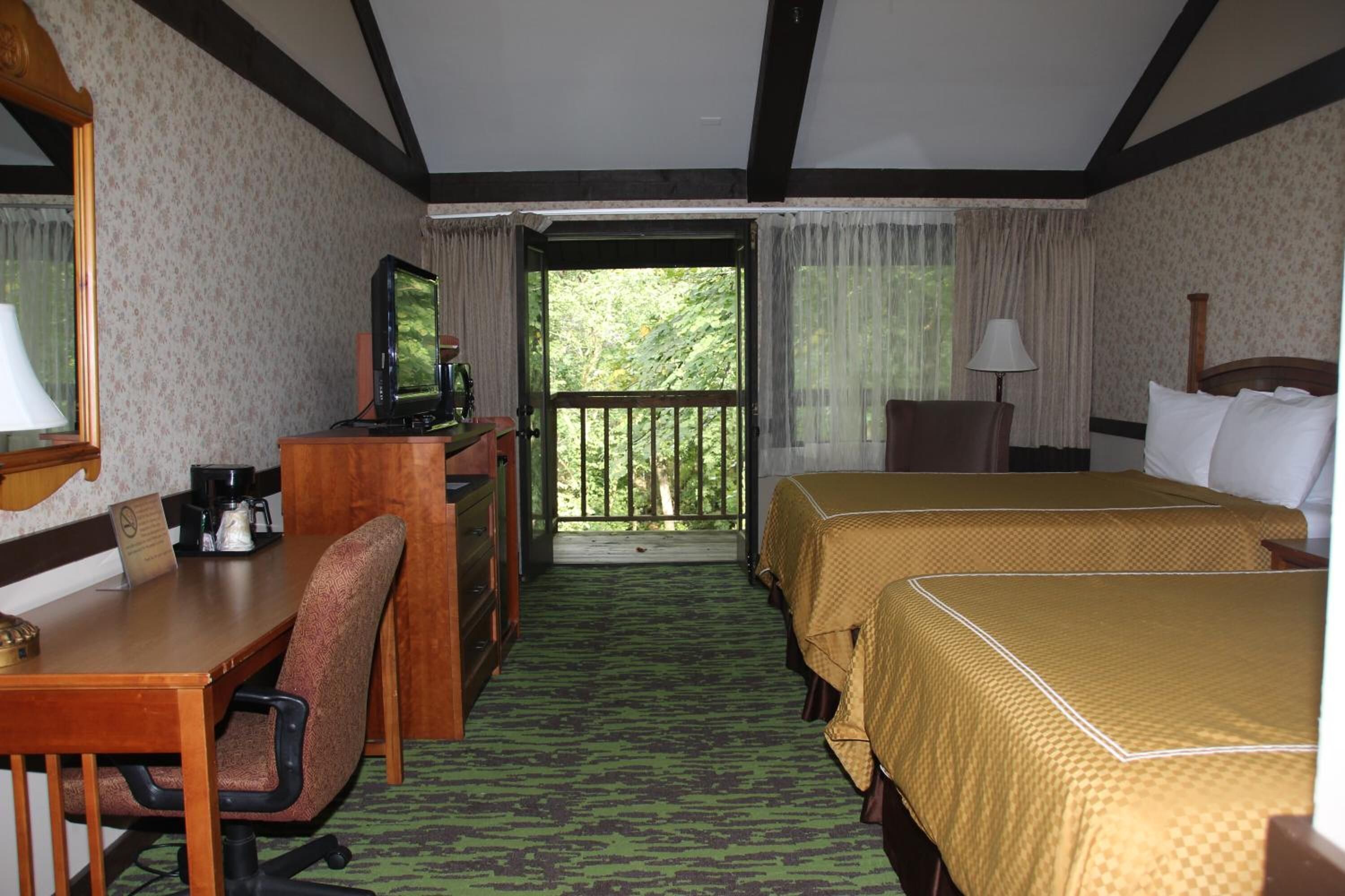 A hotel room with a desk, chair, two beds, a television, and a balcony with a view of trees.