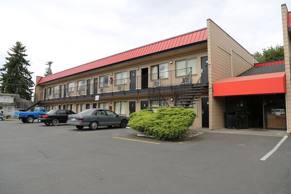 A multi-story motel with a red roof and beige walls, featuring a parking lot with several cars and a small green bush in the foreground.