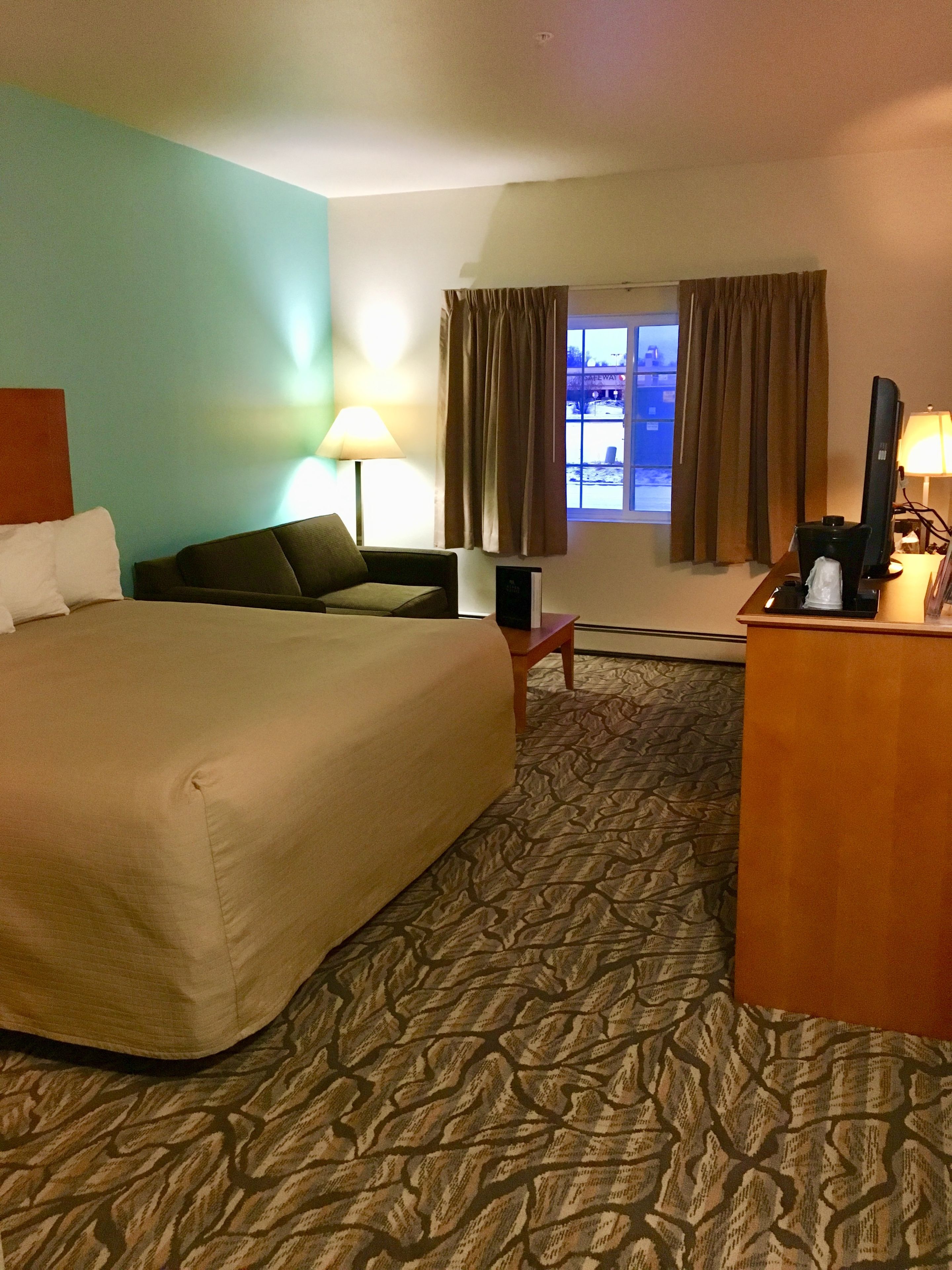 Quarto de hotel com cama, escrivaninha, cadeira, abajur e janela com cortinas.