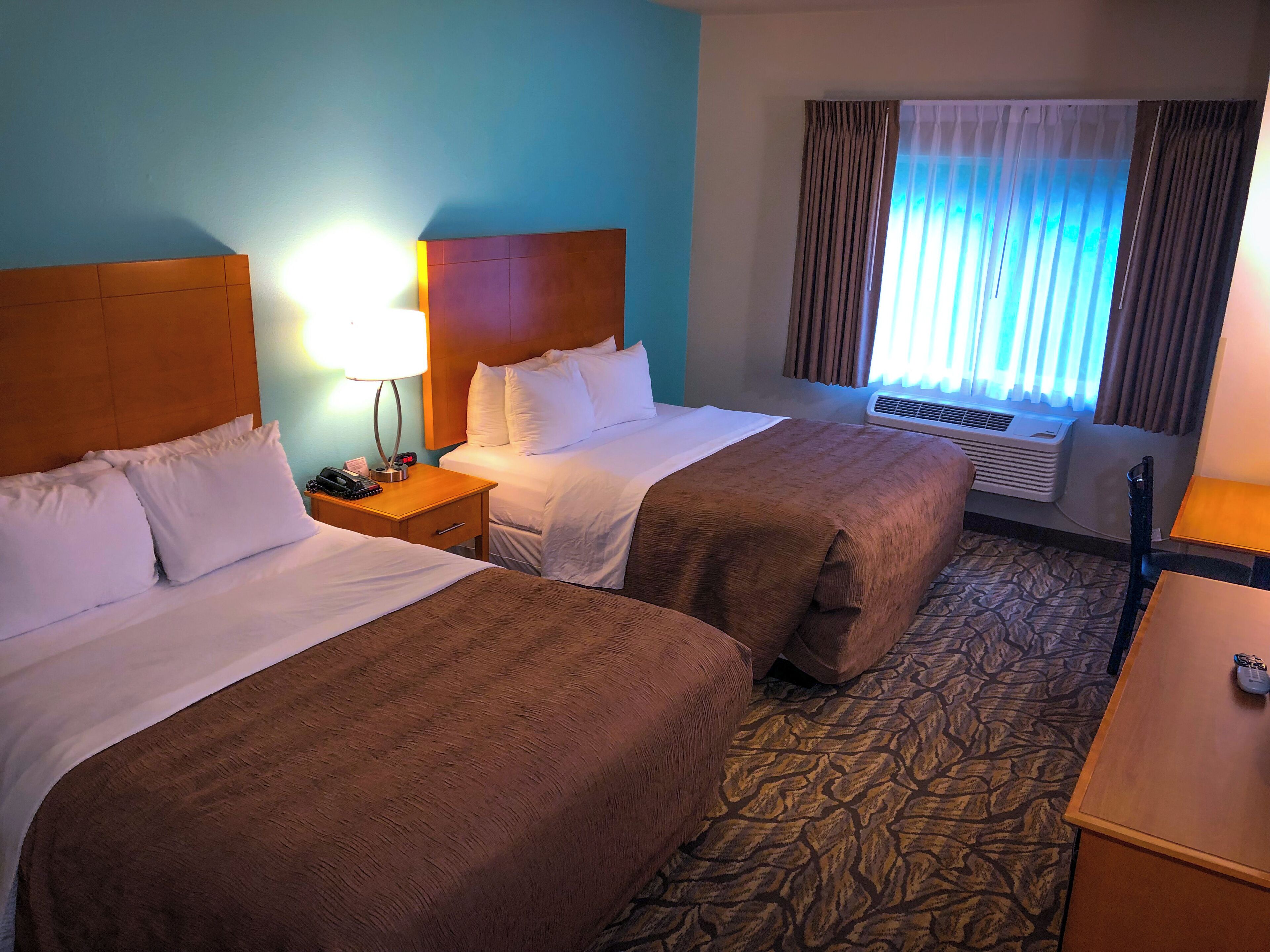 A hotel room with two beds, a nightstand with a lamp, a desk, and a window with curtains.