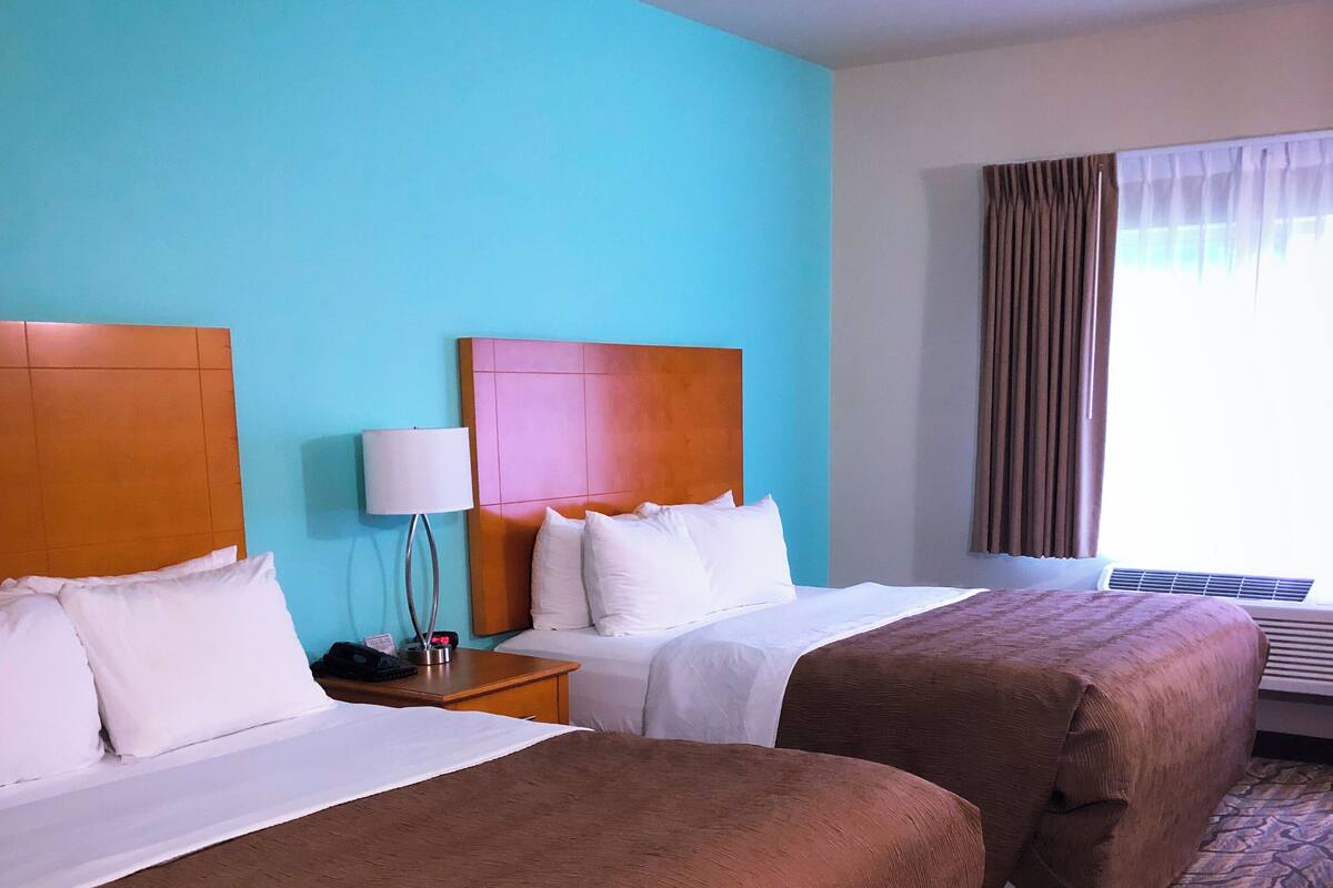 A hotel room with two beds, a nightstand, a lamp, and a window with curtains.