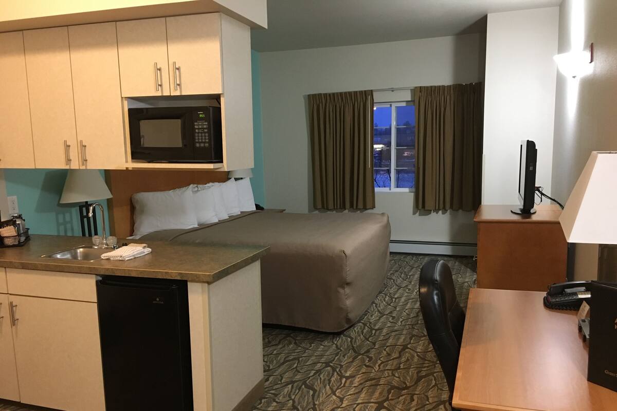 A hotel room with a kitchenette, a bed, a desk, and a window with curtains.