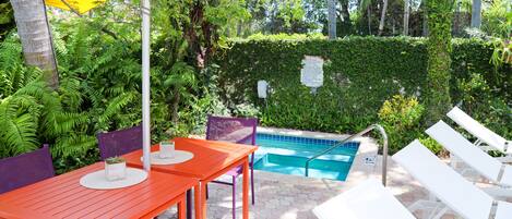 Un espace extérieur avec une piscine, une table et des chaises, le tout entouré d’une végétation luxuriante et d’une haie.