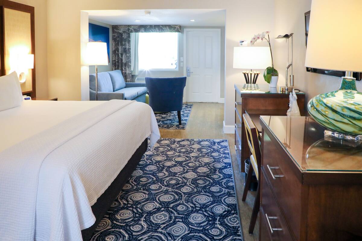 A hotel room with a large bed, a desk with a lamp, a chair, and a bathroom visible through an open door.