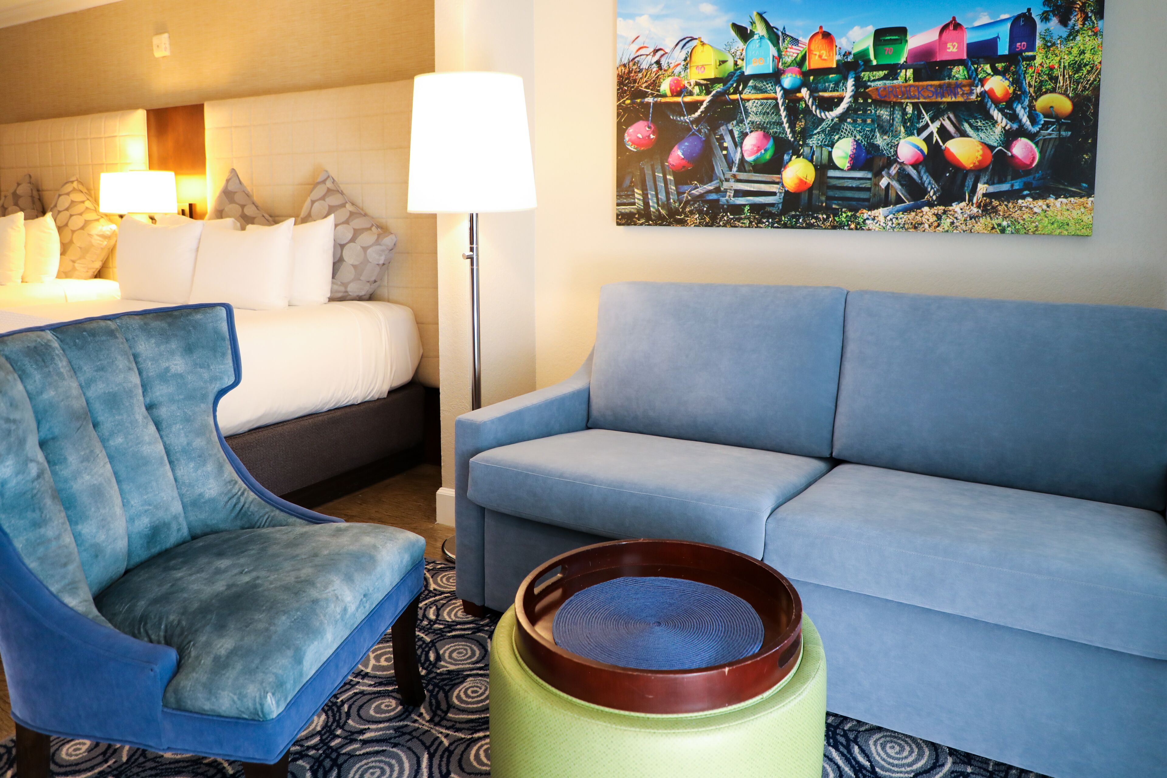 A hotel room with a bed, a blue armchair, a blue sofa, a floor lamp, and a colorful painting on the wall.