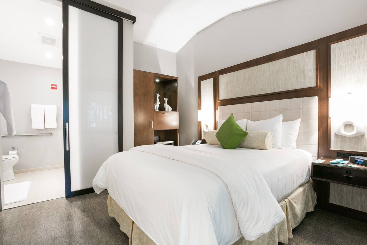 A modern hotel room with a large bed, a nightstand, a bedside table, and a bathroom visible through an open door.