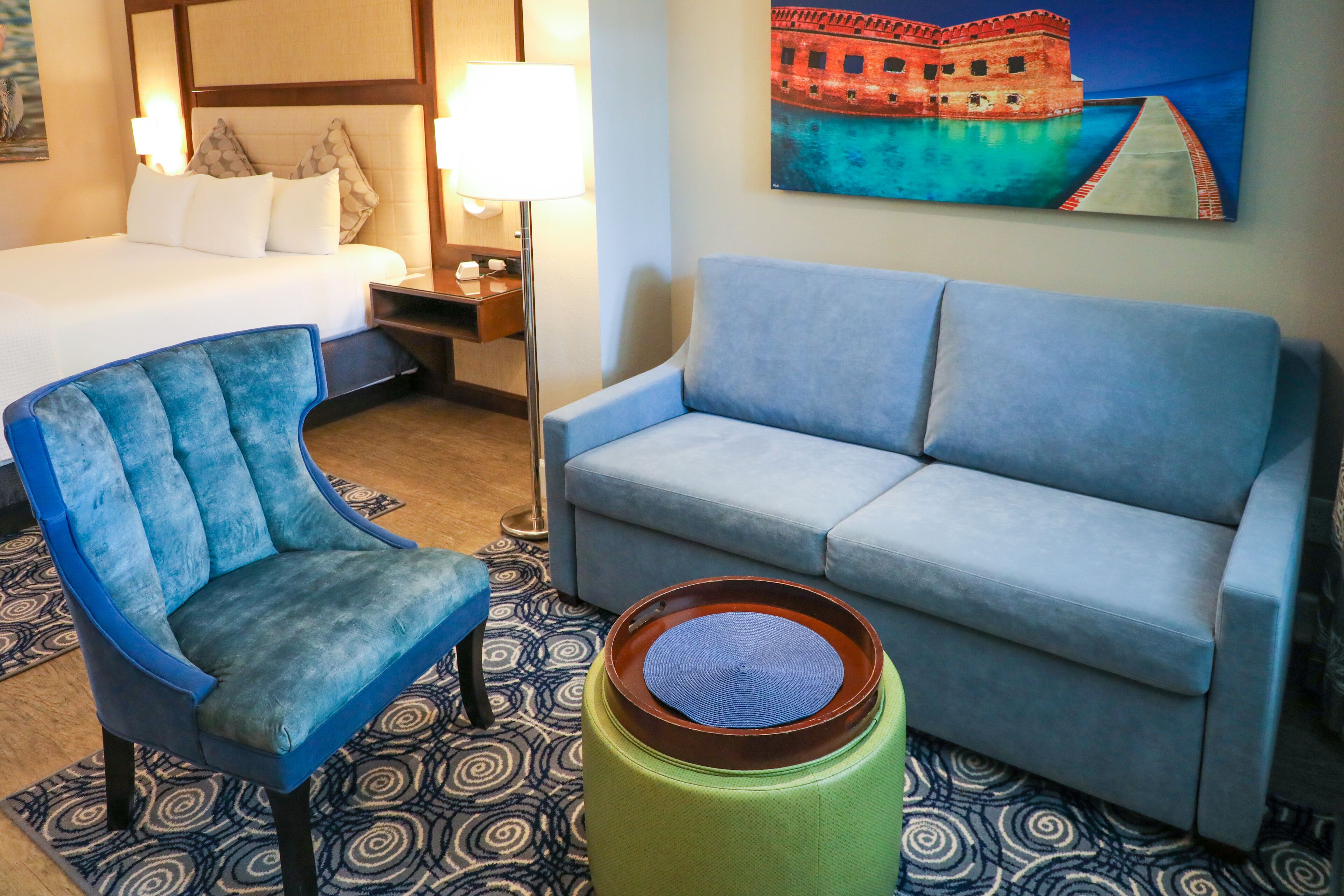 Una habitación de hotel con una cama, un sillón azul, un sofá azul, un otoman verde y un cuadro de un castillo en la pared.