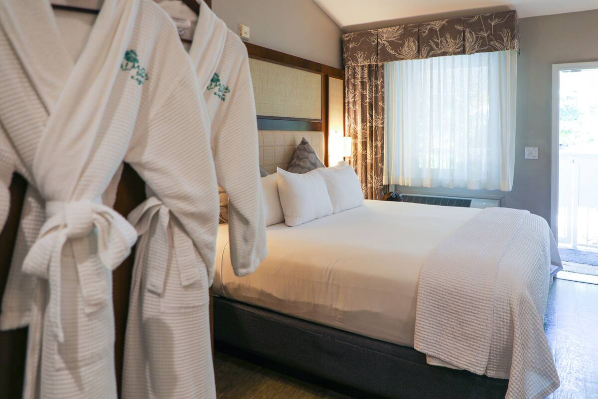 A hotel room with a large bed, a headboard, a window with curtains, and two white robes hanging on a rack.