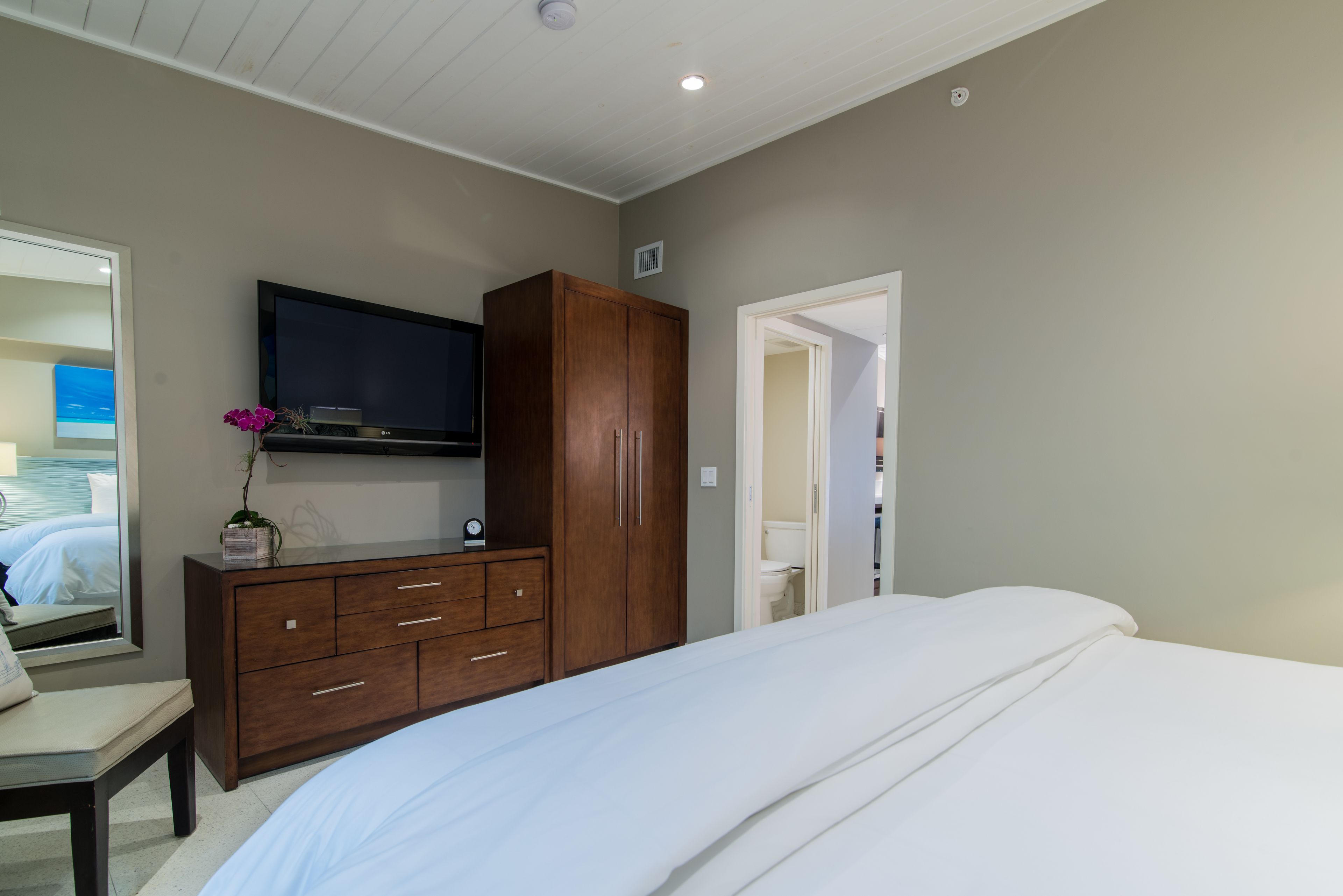 A bedroom with a bed, a wooden dresser, a flat-screen TV, and a mirror.