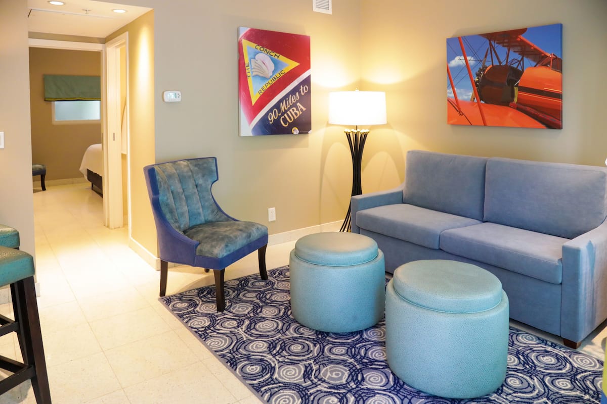 Um saguão de hotel com uma poltrona azul, um sofá azul e dois pufes sobre um tapete estampado. Há duas obras de arte na parede, uma representando um avião e outra um carro vermelho. Uma luminária de chão também é visível.
