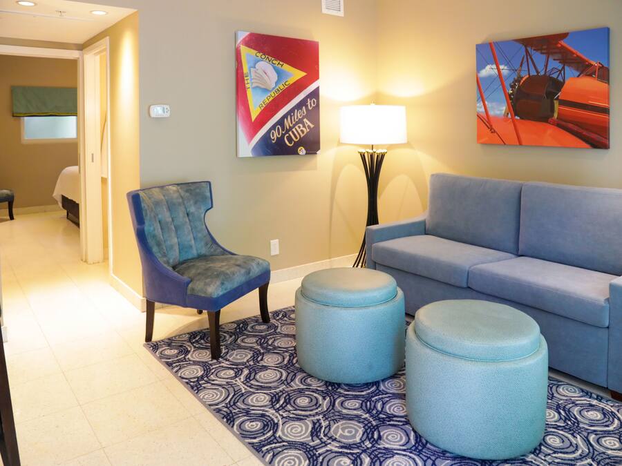 Eine Hotellobby mit einem blauen Sessel, einer blauen Couch und zwei Hocker auf einem gemusterten Teppich. Zwei Wandbilder sind zu sehen, eines zeigt ein Flugzeug und das andere ein rotes Auto. Eine Stehlampe ist ebenfalls erkennbar.