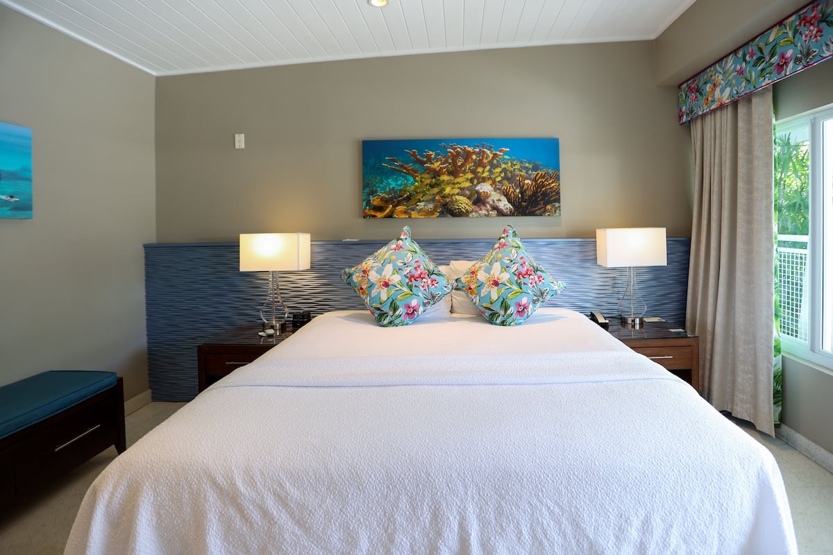 Quarto de hotel com uma cama grande, duas mesas de cabeceira com luminárias, um quadro de um recife de coral na parede e uma janela com cortinas.