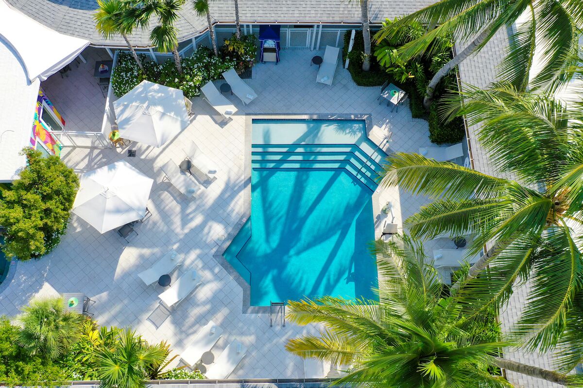 An aerial view of a hotel courtyard with a swimming pool, lounge chairs, umbrellas, and palm trees.