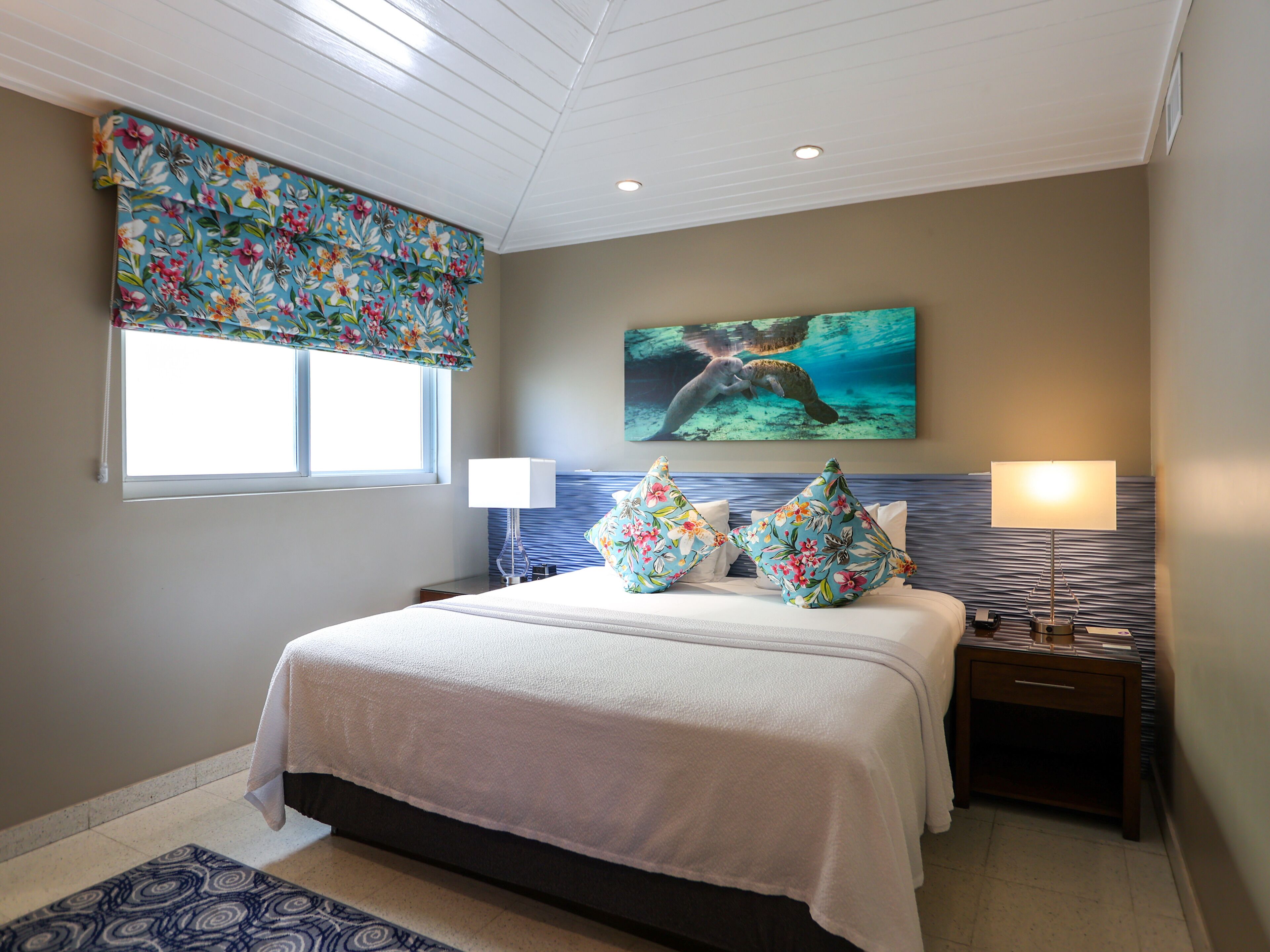 A hotel room with a bed, two bedside tables, a window with a floral curtain, a wall-mounted painting of a sea turtle, and a patterned rug.