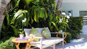 A patio with a white wicker lounge chair, a wooden table with a book and a potted plant, surrounded by lush tropical foliage and palm trees. - Orchid Key Inn - Adults Only (Key West)