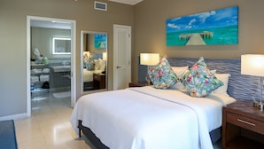 A hotel room with a large bed, a nightstand, a desk, a bathroom with a shower, and a painting of a pier over water on the wall. - Orchid Key Inn - Adults Only (Key West)