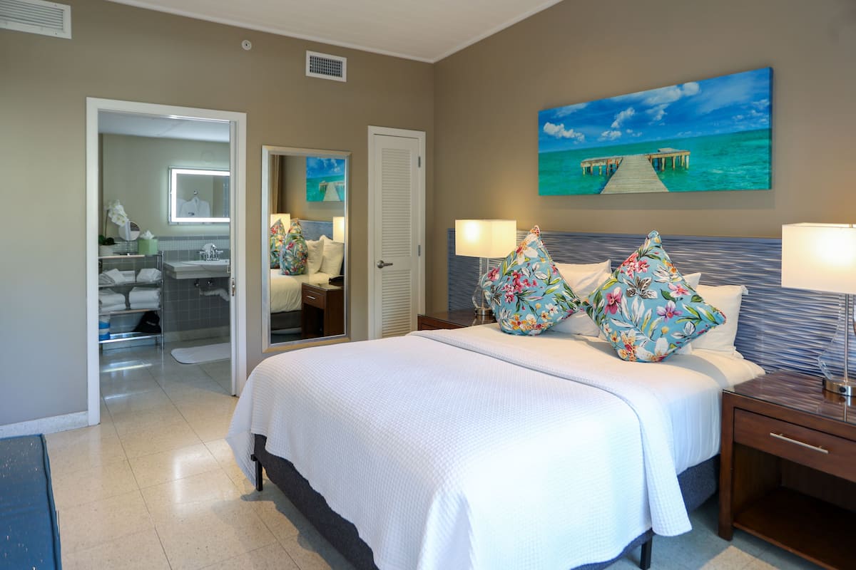 Quarto de hotel com uma cama grande, mesa de cabeceira, escrivaninha, banheiro com chuveiro e um quadro de um cais sobre a água na parede.