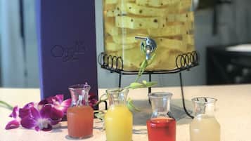 Un dispensador de bebidas con un tarro de vidrio lleno de rodajas de fruta, rodeado de pequeñas botellas de vidrio con bebidas de diferentes colores, y una caja morada con la etiqueta "Orchid Bar" al fondo.