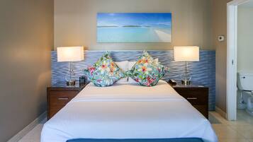 A hotel room with a bed, two nightstands, a painting of a beach, and a bathroom visible through an open door.
