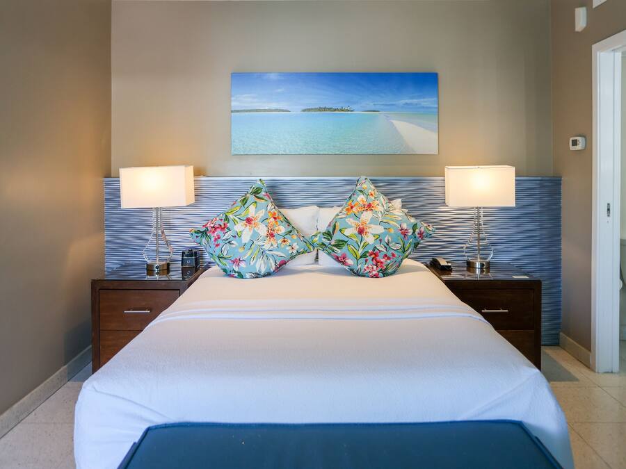 A hotel room with a bed, two nightstands, a painting of a beach, and a bathroom visible through an open door.