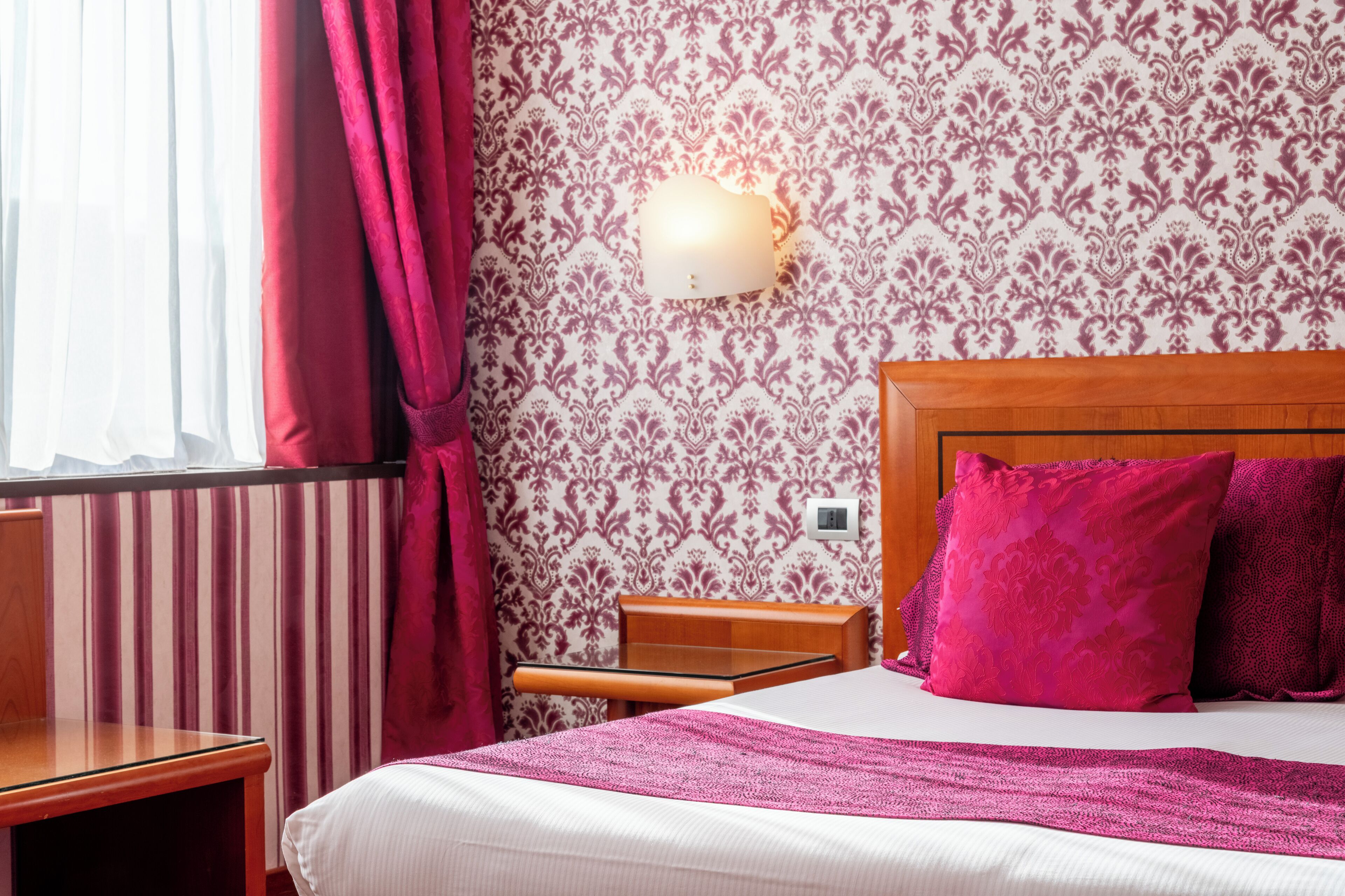 Una habitación de hotel con cama, mesita de noche, lámpara y ventana con cortinas.