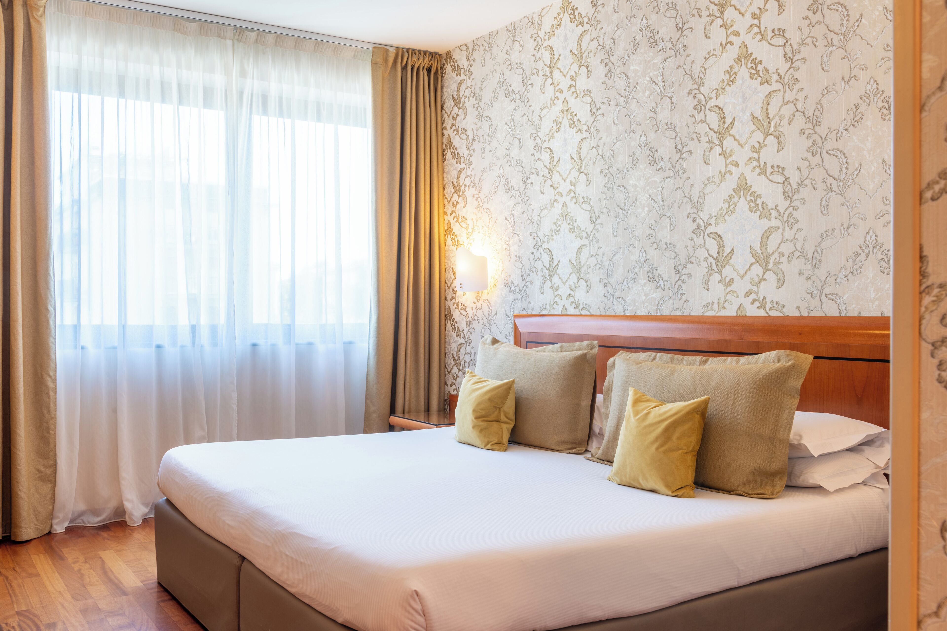 A neatly made bed with pillows, a wooden headboard, and a window with sheer curtains.