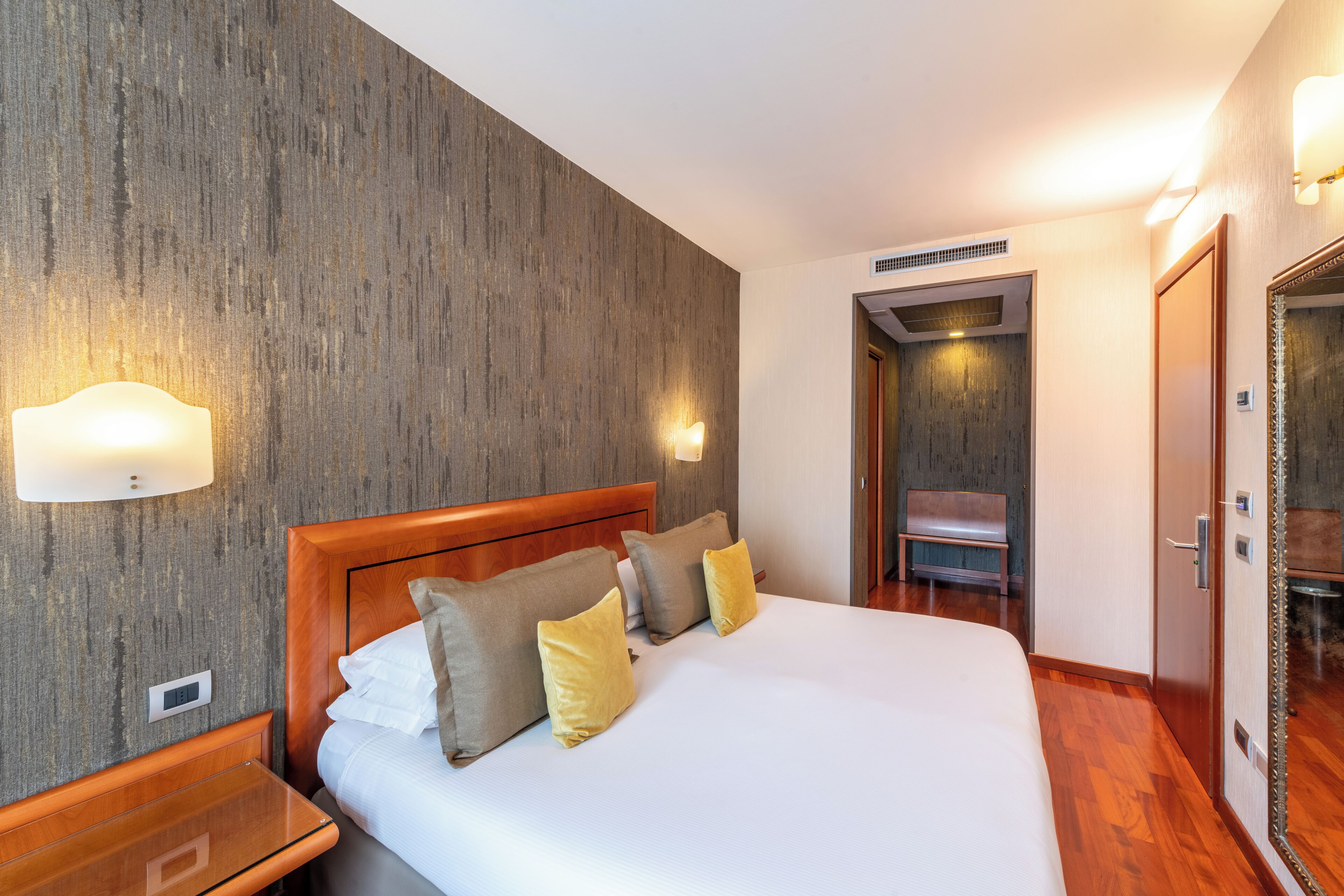 Ein Hotelzimmer mit einem hölzernen Kopfteil, einem Bett mit weißen Leinentüchern und mehreren Kissen, einem Nachttisch, einer Bank und einem Spiegel.