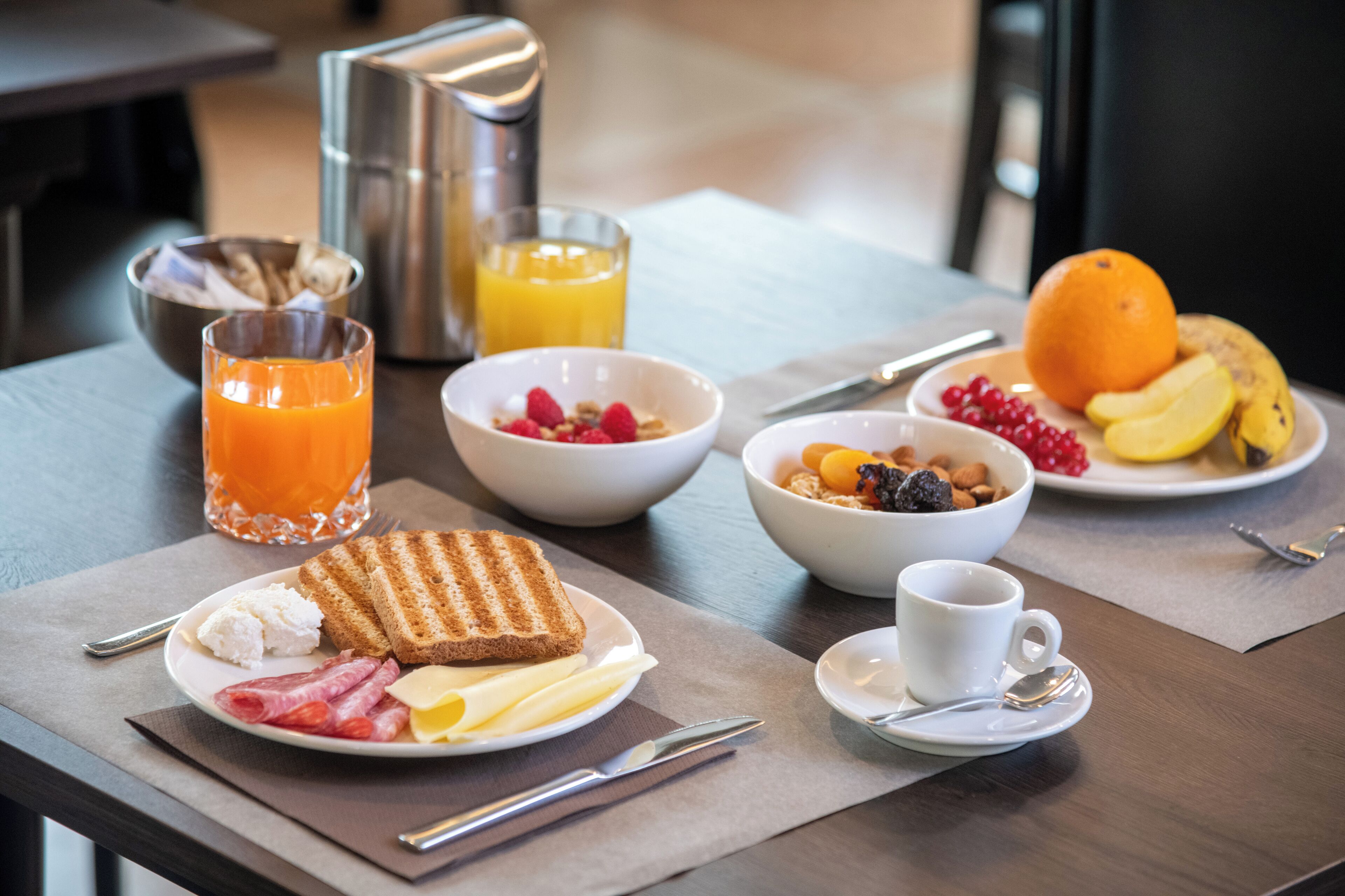 A breakfast setting with toast, eggs, bacon, and fruit on plates, a bowl of cereal, a glass of orange juice, and a coffee cup.