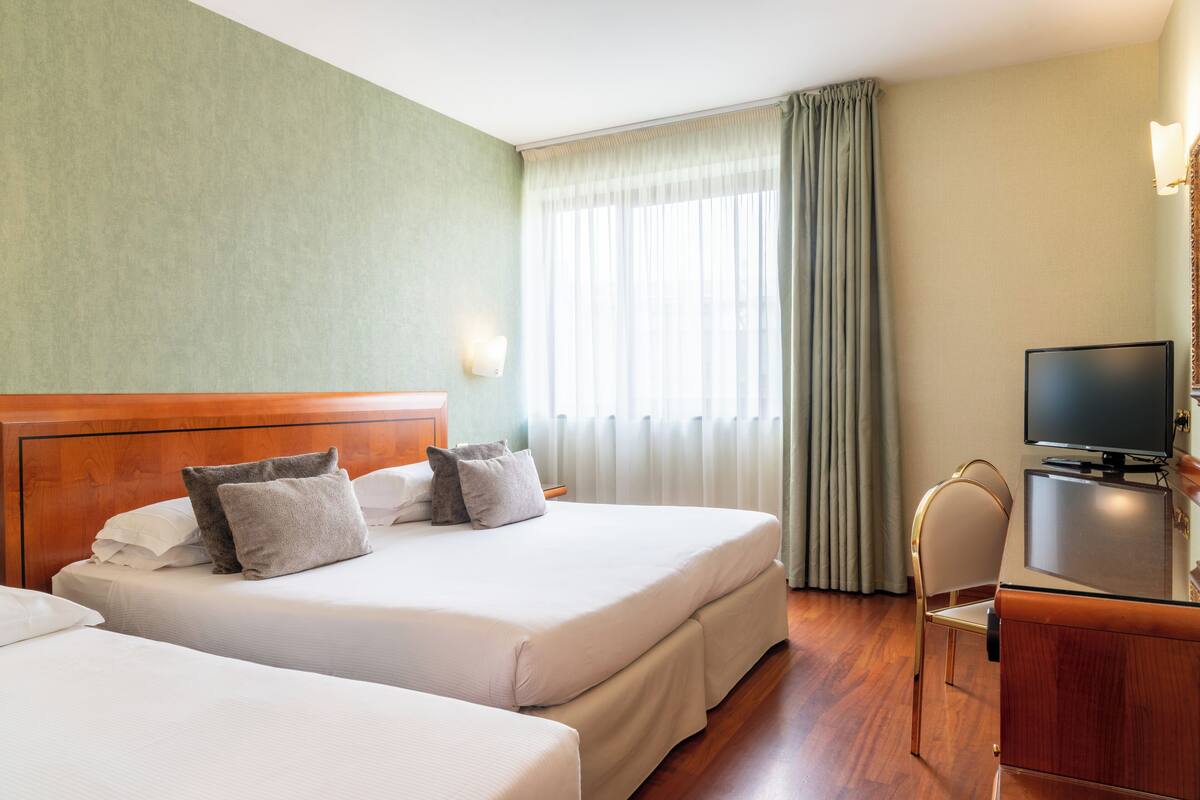 A hotel room with two beds, a desk, a television, and a window with curtains.
