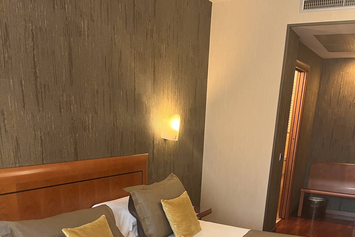 A hotel room with a wooden headboard, a bed with grey and gold bedding, a grey throw blanket, and a wall-mounted light fixture.
