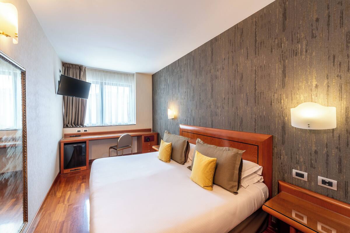 A hotel room with a large bed, a desk with a computer, a TV, and a window with curtains.
