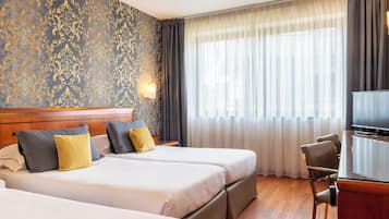 A hotel room with two beds, a TV, a desk, and a large window with curtains.
