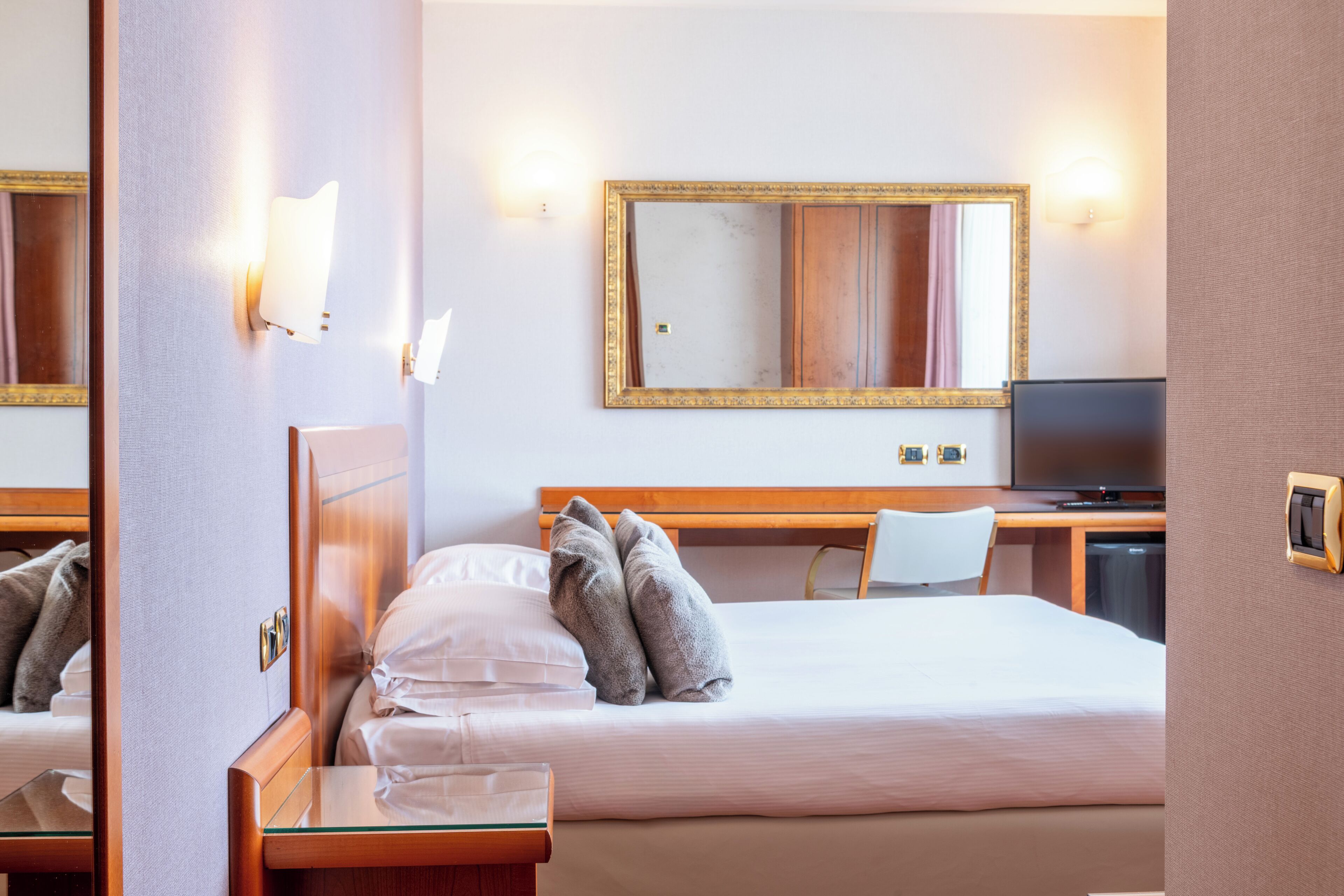 A hotel room with a bed, a desk with a chair, a television, and a large mirror on the wall.