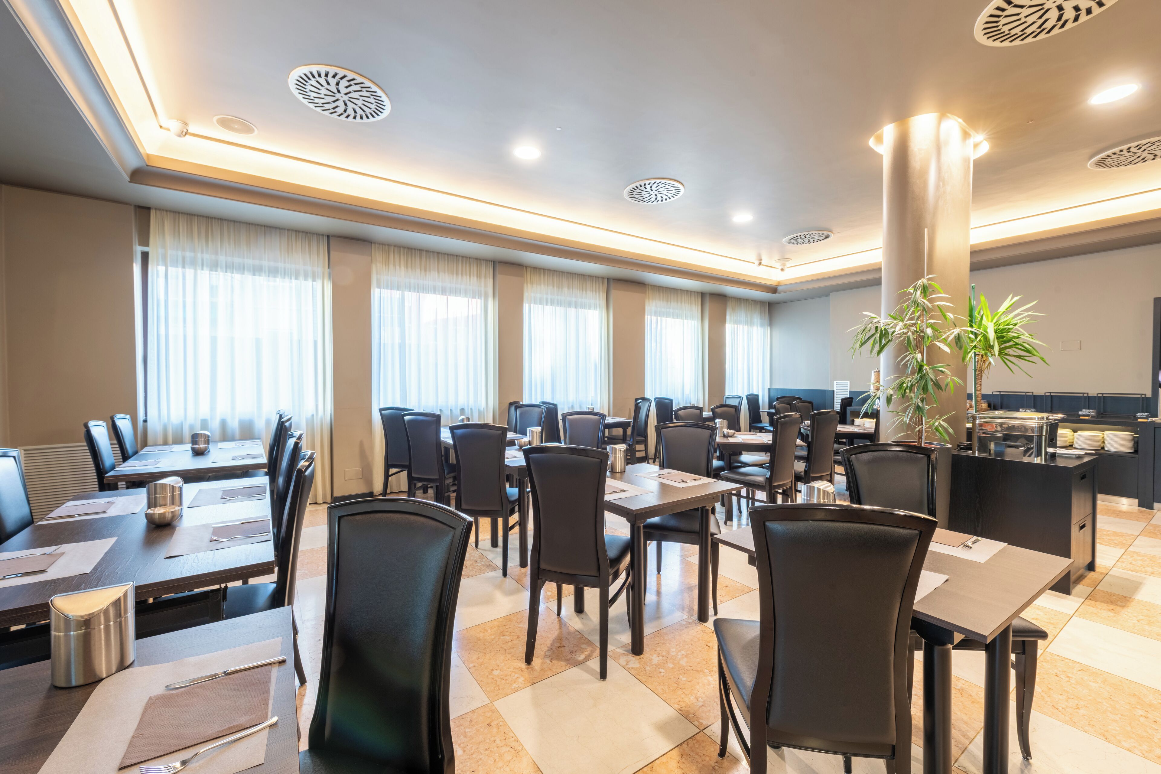 Comedor de hotel con varias mesas y sillas, zona de cocina y un ventanal con cortinas.