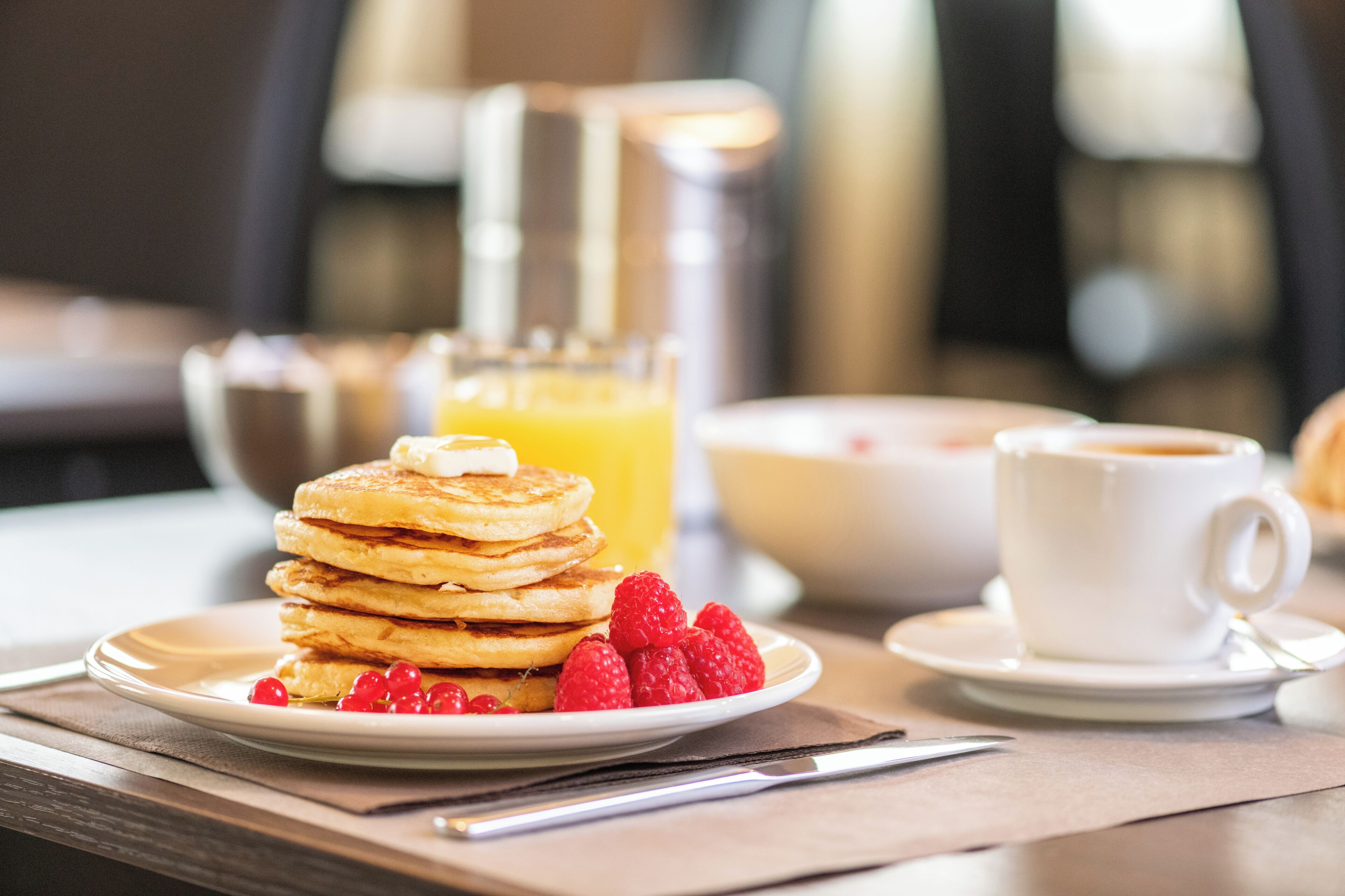 A breakfast setting with pancakes, raspberries, a glass of orange juice, and a cup of coffee on a table.