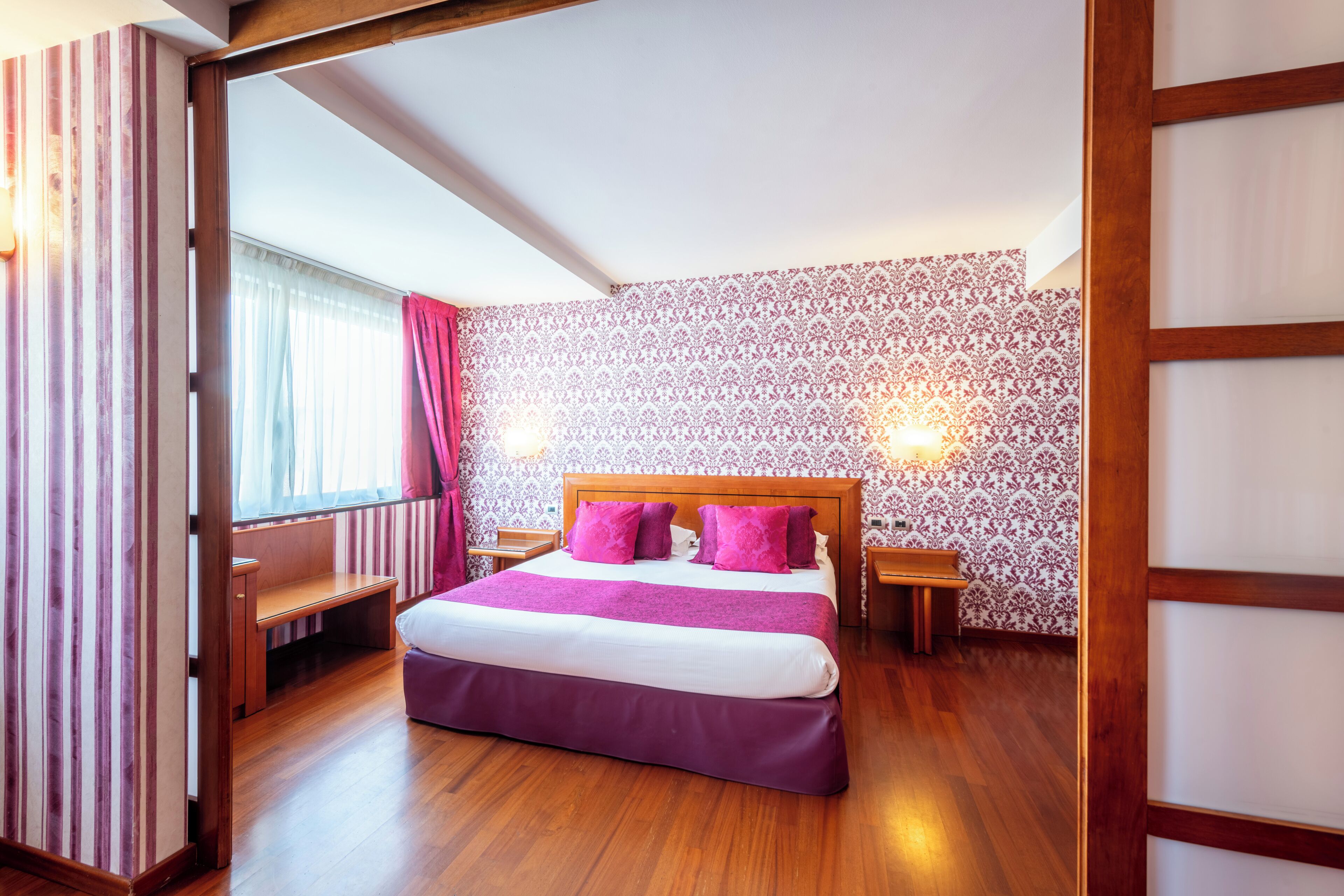 Un dormitorio con una cama grande, cabecera de madera, dos mesitas de noche, un banco y papel tapiz estampado.