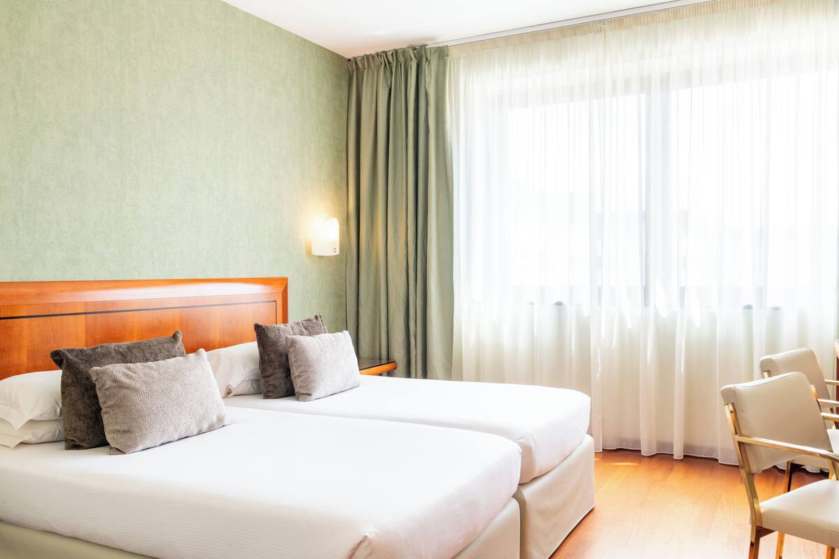 A hotel room with two beds, a wooden headboard, a nightstand, a lamp, a chair, and a large window with curtains.