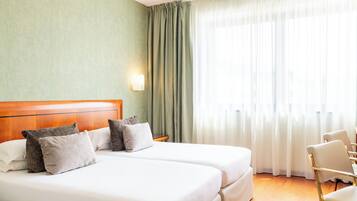 Habitació d'hotel amb dos llits, un capçal de fusta, una taula de nit, una làmpada, una cadira i una finestra gran amb cortines.