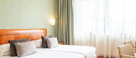 A hotel room with two beds, a wooden headboard, a nightstand, a lamp, a chair, and a large window with curtains.