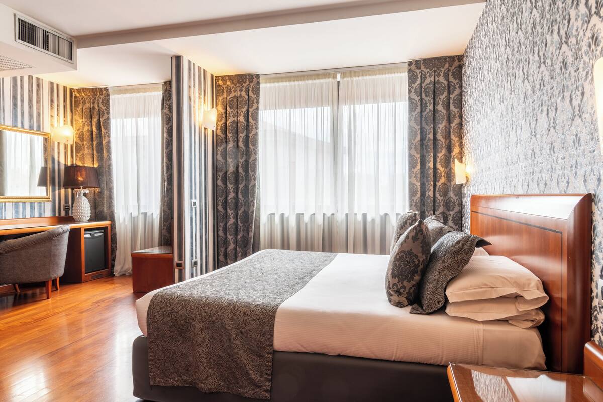 A hotel room with a large bed, a desk, and a chair. There is a window with curtains and a wall with a patterned wallpaper.
