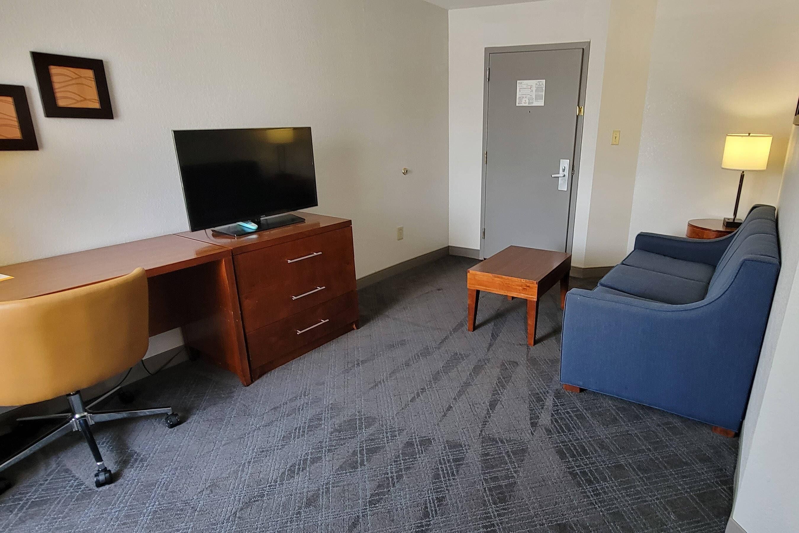 A hotel room with a wooden desk, a television, a blue sofa, a small table, and a lamp.