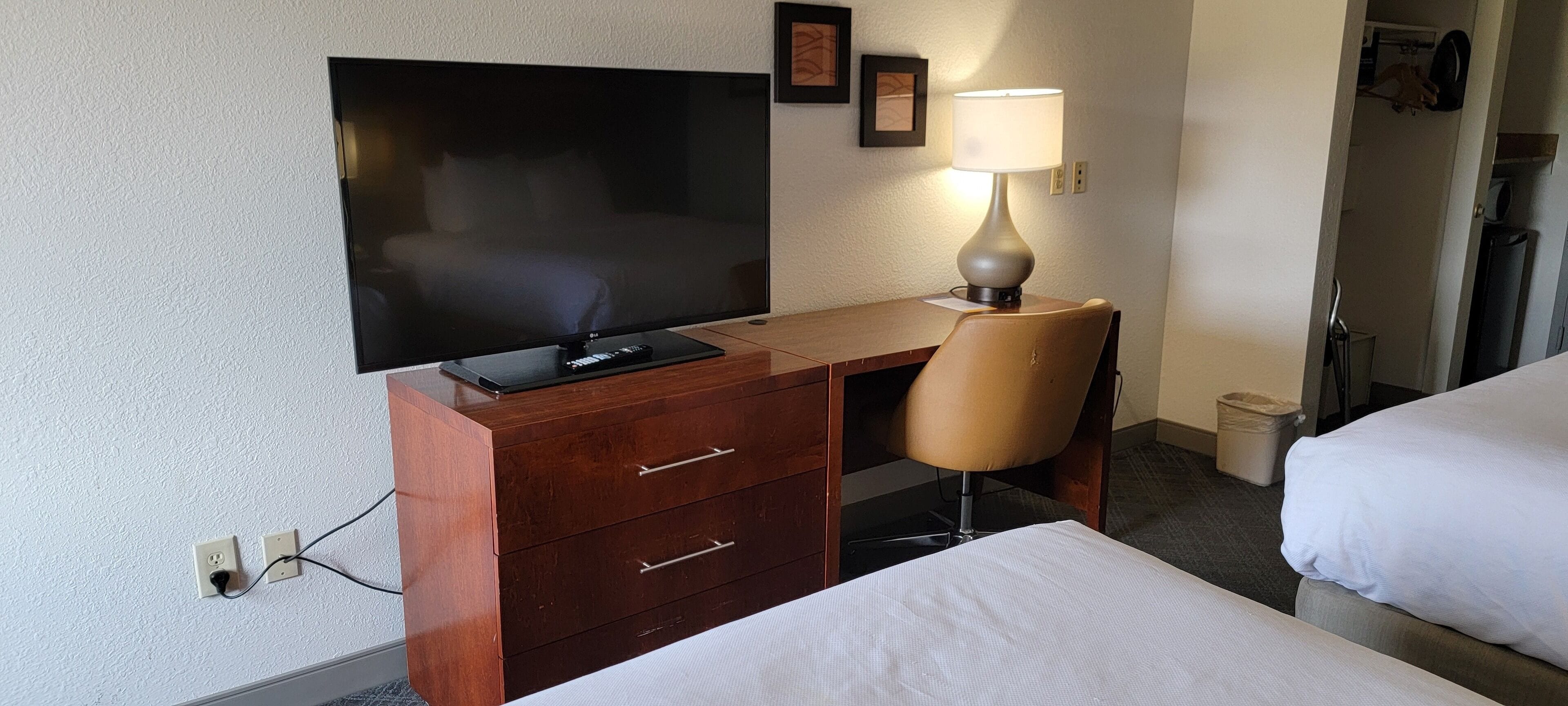 A hotel room with a TV, a desk with drawers, a chair, and a lamp.