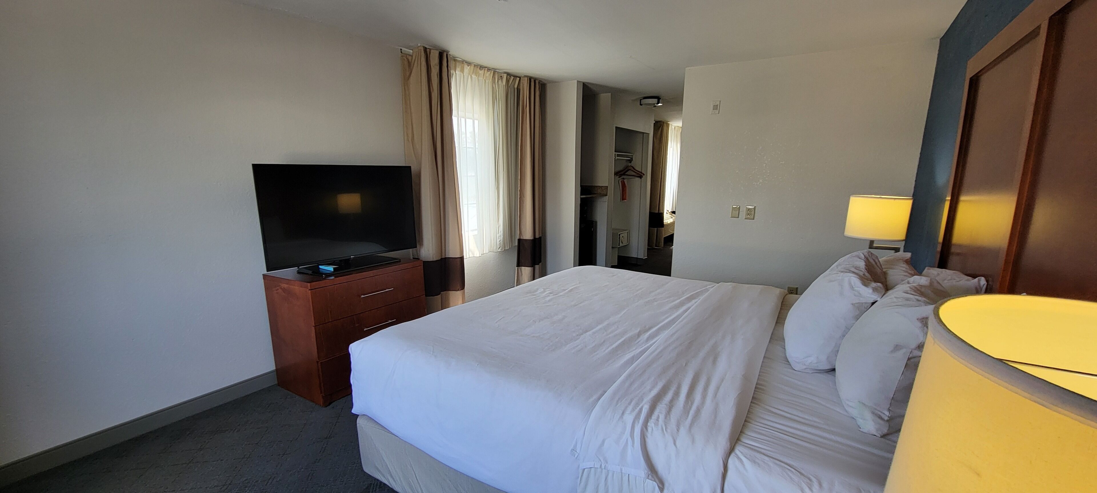 A hotel room with a bed, a television on a wooden dresser, a lamp, and a door leading to a bathroom.