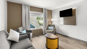 A modern hotel room with a grey sofa, a small armchair, a yellow side table, a flat-screen TV mounted on the wall, and a window with curtains.